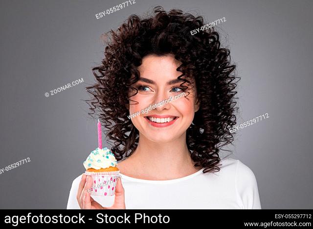Cheerful beautiful curly young woman making a wish holding birthday cupcake with candle