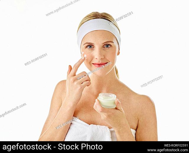 Woman wearing headband and towel applying face cream while standing against white background