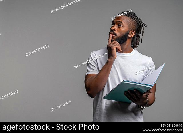 Waist-up portrait of a pensive young man with an open notebook standing by the gray wall