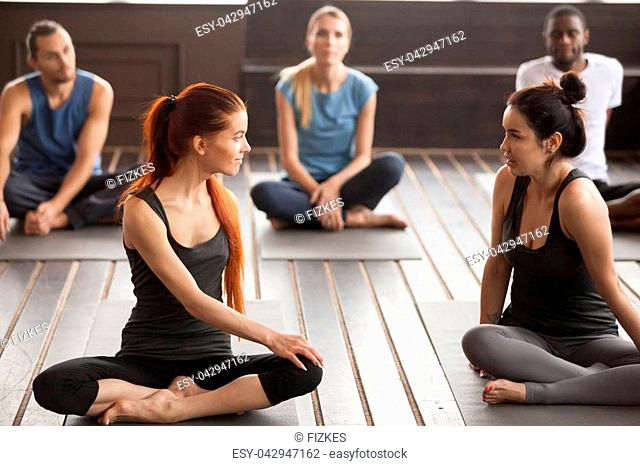 Two young beautiful fit yogi women talking at group training class, pilates instructors trainers discussing yoga exercises before session sitting on mats in gym...
