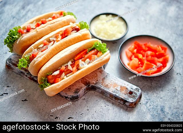 Three barbecue grilled hot dogs with sausage placed on wooden cutting board. Bowls with tomato and onionon sides. Traditional american fast food
