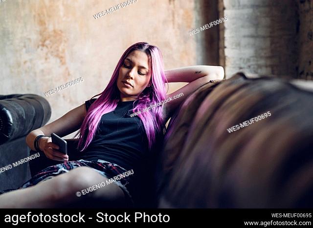 Young woman with pink hair sitting on sofa in a loft using smartphone