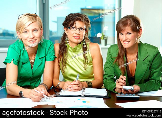 Young businesswomen sitting at table in meeting room, discussing charts on table, looking at camera, smiling
