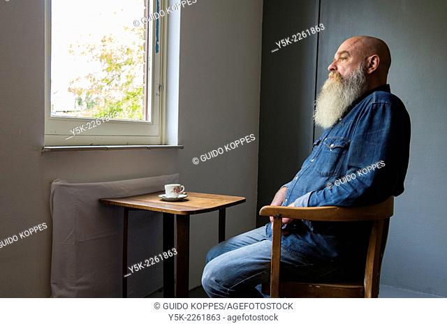 Tilburg, Netherlands. Portrait of a bald man with beard, drinking a cup of coffee while looking out the window