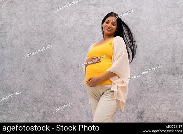 Happy pregnant woman standing against wall with holding her stomach