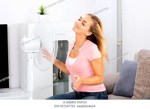 Young Smiling Woman Sitting On Couch In Front Of Fan During Hot Weather At Home