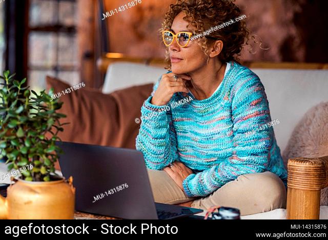 Serene pretty adult woman at home after online use of laptop computer sitting on the sofa and looking outside. Happy modern female peoplerelax indoor in living...