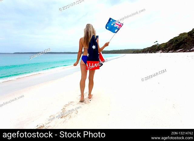 Aussie blonde woman walking along an idyllic beach wearing a bikini with patriotic Australian flag towel draped across one shoulder