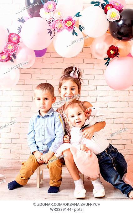 Boys and girl with crowns under birthday balloon and paper flower arch decorations. Childish photozone for celebration