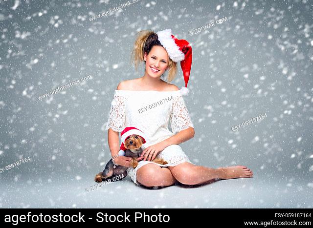 Beautiful happy girl in lace dress and red santa cap sitting with pretty yourkshire terrier in christmas hat. Studio shot over grey background with falling snow