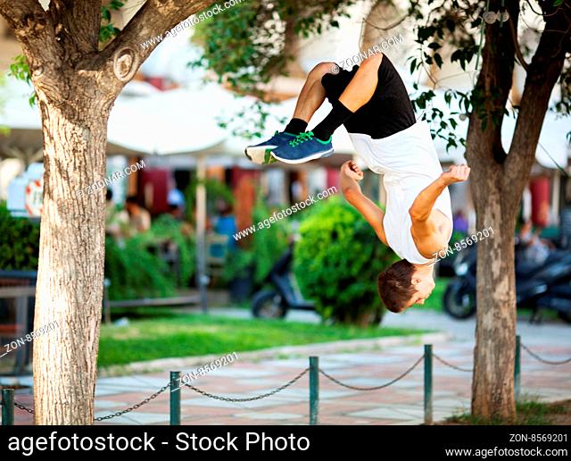 Young extreme athlete doing front flip between the trees. City street with outdoor cafe in background