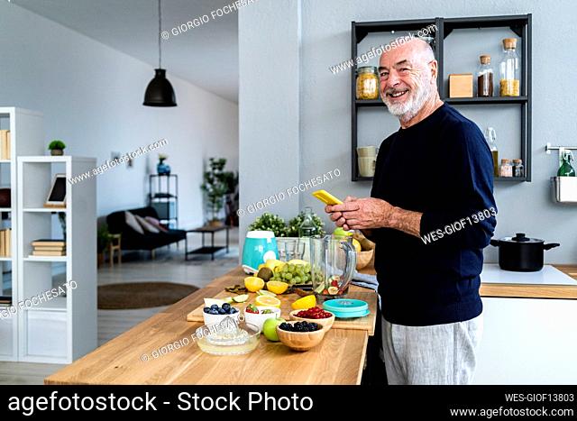 Smiling man with mobile phone standing at kitchen counter