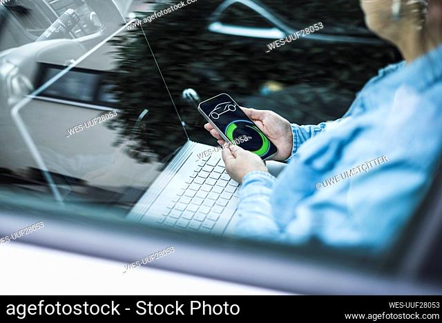 Mature woman with laptop and electric car charging app on mobile screen seen through glass