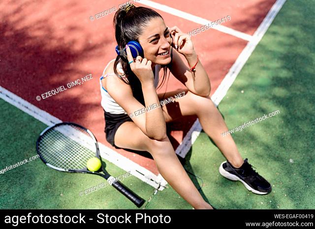 Smiling female tennis player listening music while sitting on floor in court
