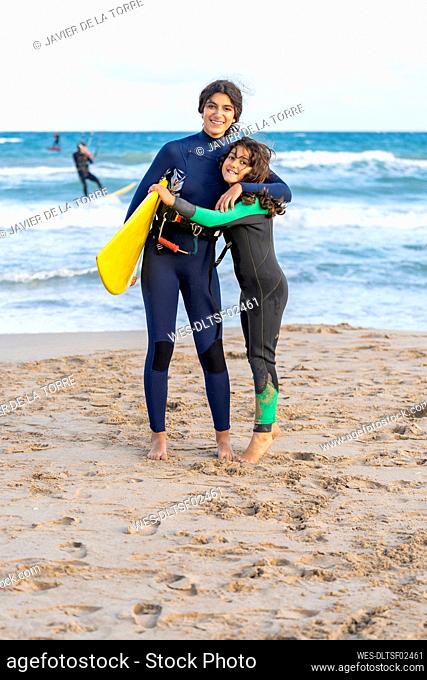 Smiling girl with kiteboard embracing sibling on sand at beach