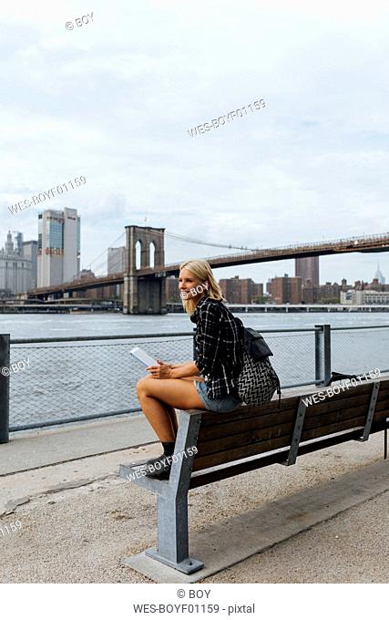 USA, New York City, Brooklyn, smiling young woman sitting at the waterfront with backpack and tablet