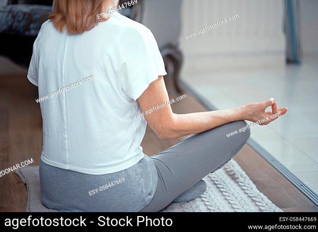 Woman In Casual Cloth Sits With Crossed Legs In The Lotus Pose Meditating. Sitting In Asana While Practicing Yoga. Back View. Selective Shot