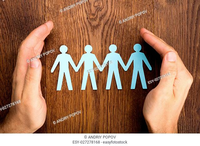 Close-up Of Person Hand Protecting Human Figure Cutout On Wooden Desk