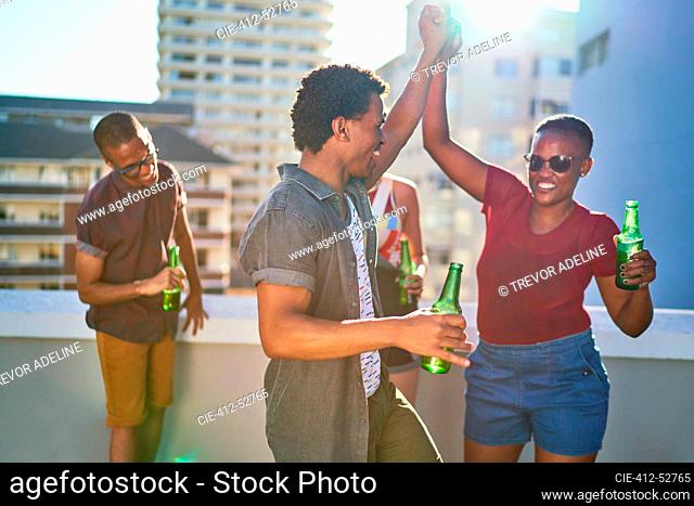 Happy young friends dancing and drinking beer on sunny urban rooftop