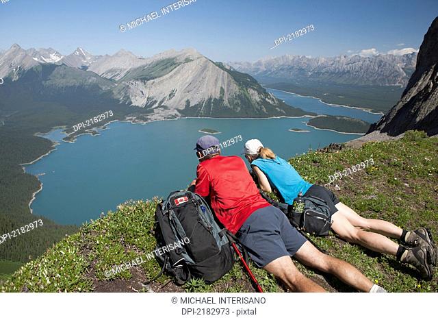 Male And Female Hiker Laying Down On A Mountain Ridge Overlooking An Emerald Lake And Mountains Below With Blue Sky In Kananaskis Provincial Park