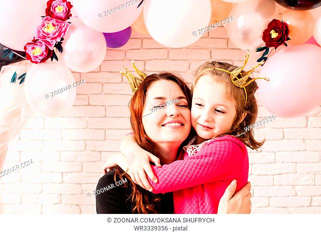 Two beautiful girls with crowns under birthday balloon and paper flower arch decorations. Childish photozone for celebration. Mom with dauther