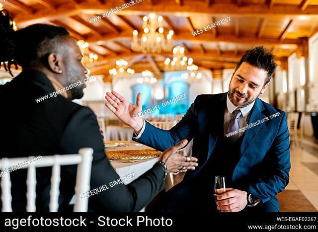 Male friends greeting while sitting and holding champagne at banquet