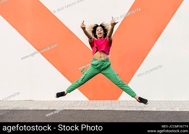 Cheerful woman making obscene hand gesture jumping in front of wall