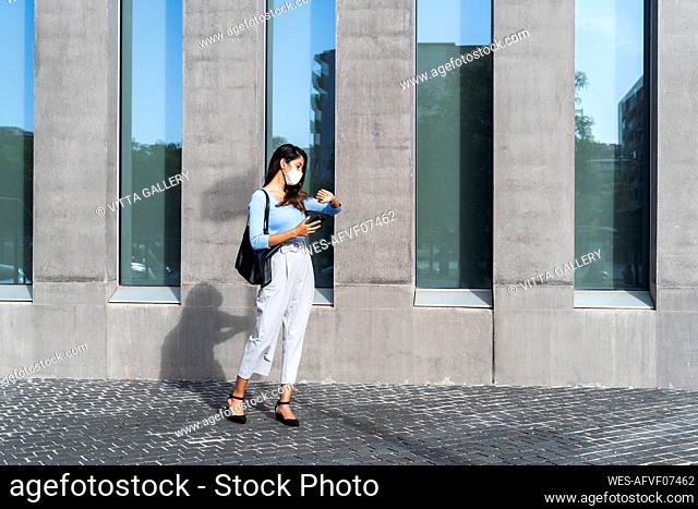 Businesswoman in face mask checking time on wristwatch while standing on footpath during pandemic