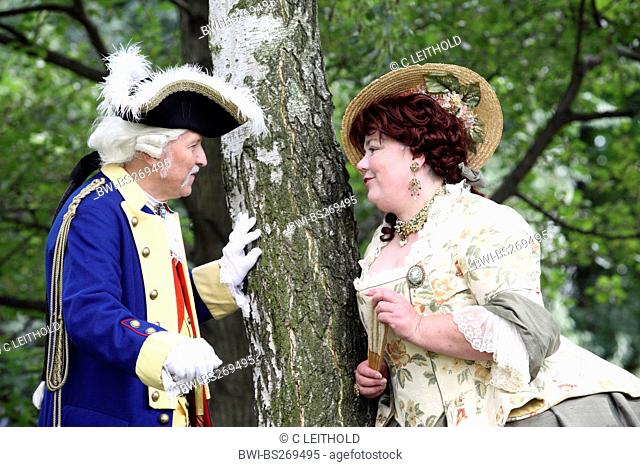older couple wearing baroque clothing standing at a garden tree flirting