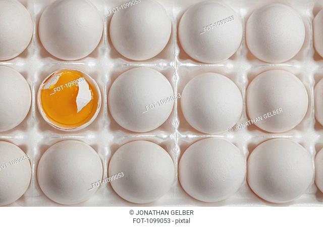 A broken egg and its egg yolk amongst eggs in a carton