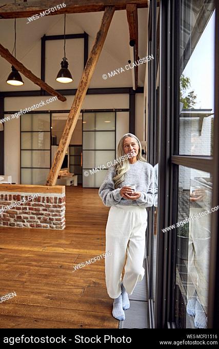 Mature woman with coffee cup day dreaming while leaning on glass door at home