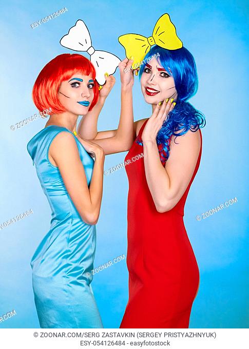 Portrait of young women in comic pop art make-up style. Females in red and blue wigs on blue background. Girls with yellow bow-tie in hands