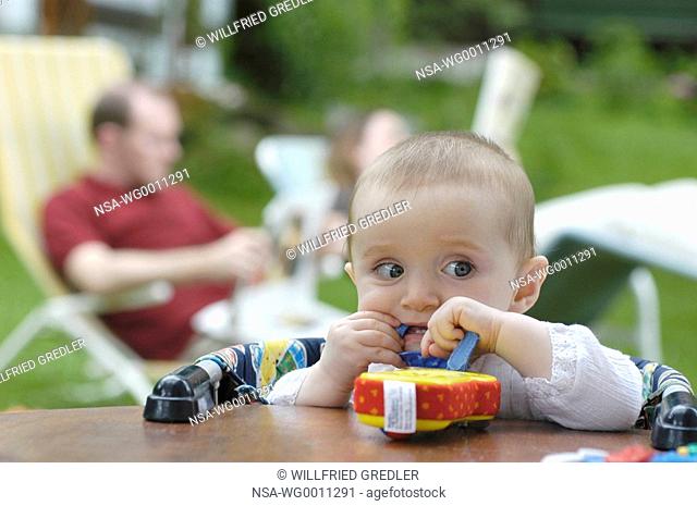 Baby plays at a tabel in the garden and parents reading newspaper