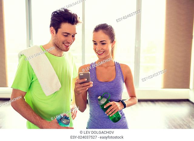 fitness, sport, technology and slimming concept - smiling young woman and personal trainer with smartphone and water bottles in gym