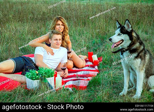 Beautiful young happy loving couple on picnic lying down on plaid with their dog in field on sunny summer day enjoying, holding and drinking wine and resting
