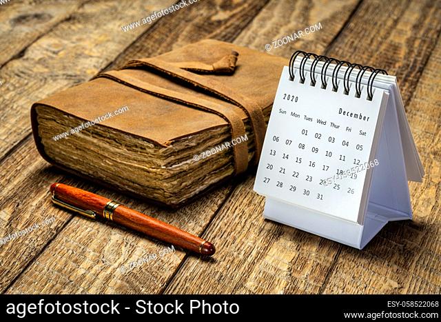 December 2020 - spiral desktop calendar against rustic wood with an antique leather-bound journal and a stylish pen, season, time and business concept