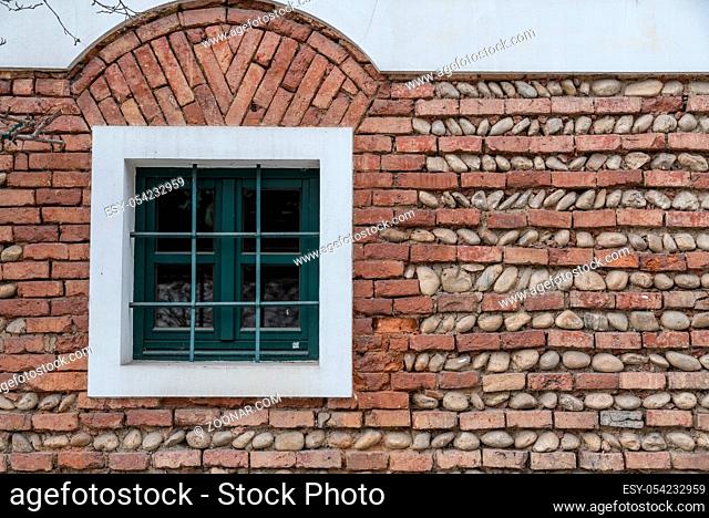 grünes Fenster mit Fenstergitter inmitten einer Ziegelmauer aus Backsteinen und Flusssteinen
