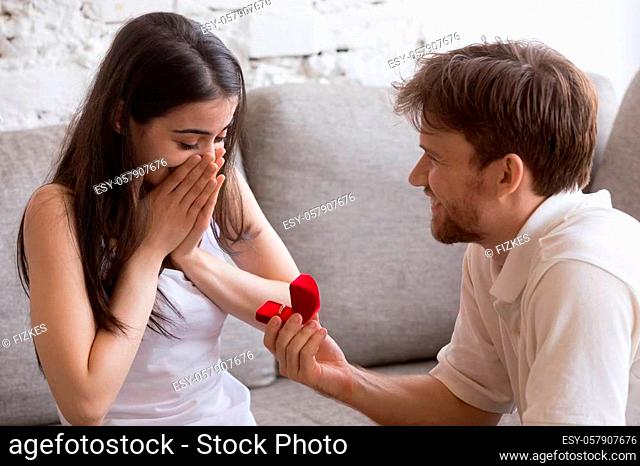 Young couple in love sitting at couch at home. Smiling man holding red box with marriage engagement ring got up on his knee and make proposal to happy surprised...