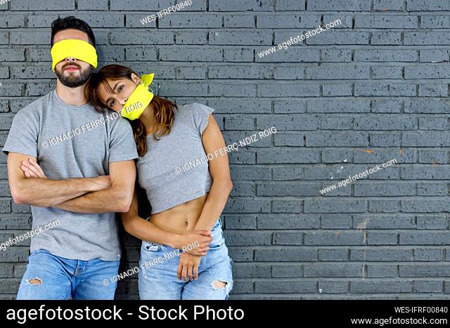 Girlfriend leaning on blindfolded boyfriend in front of gray wall