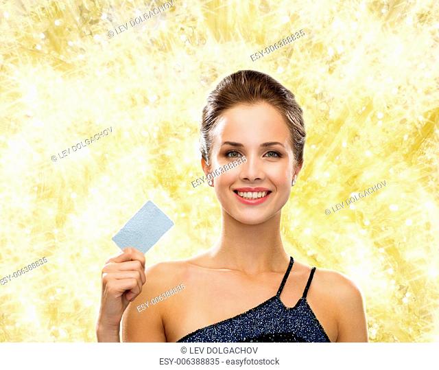 shopping, wealth, christmas, holidays and people concept - smiling woman in evening dress holding credit card over yellow lights background