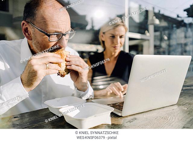 Businessman eating hamburger in coffee shop, while colleague is using laptop