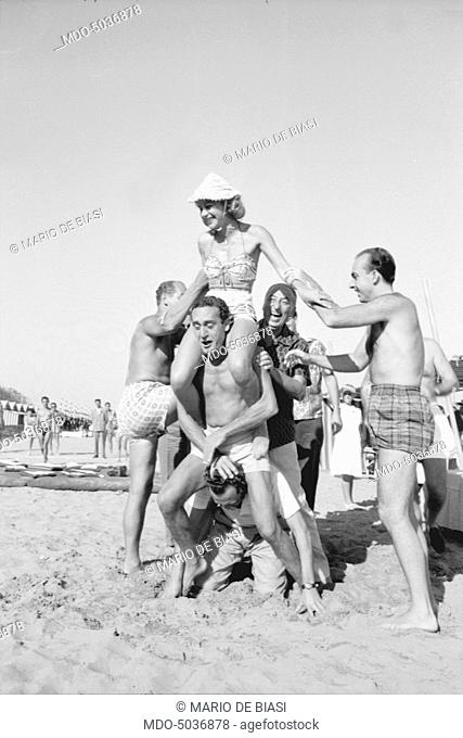 A man holding a woman on his back on the beach during the XVIII Venice International Film Festival. Venice, 1957