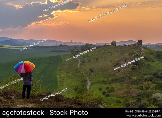 14 May 2021, Saxony-Anhalt, Thale: A hiker watches the sunset at the Teufelsmauer in the Harz mountains. He has opened an umbrella in the colours of a rainbow