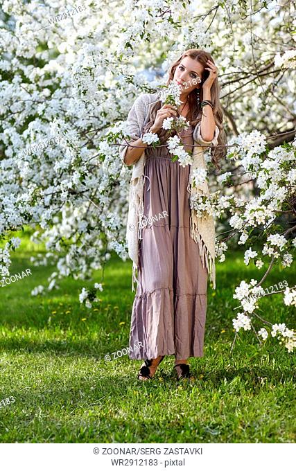 Beautiful young woman in long dress boho style on green grass under apple tree in blossom in garden