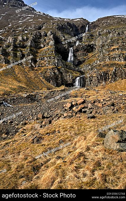 Stream flowing on cascade between rocks with remains of snow on the background of the blue sky with clouds in Iceland. In front of the rocks there is a field...