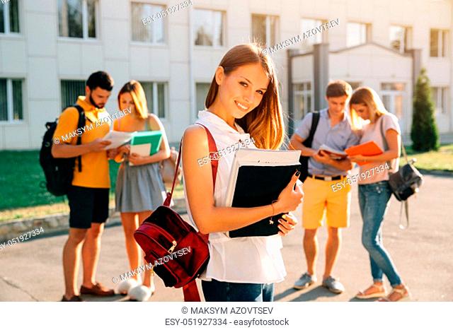 Student. Enjoying university life. Handsome young girl with red velvet backpack holding books and smiling while standing against university with her friends in...