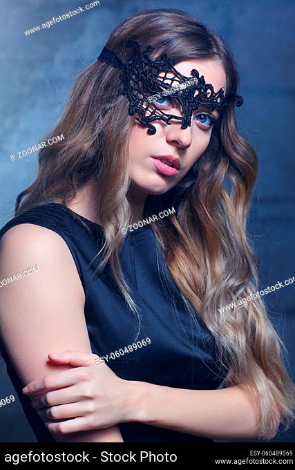 Young woman in black lace mask and black dress. Blonde female with long hair and hand on shoulder. Female on smoke and metal wall background