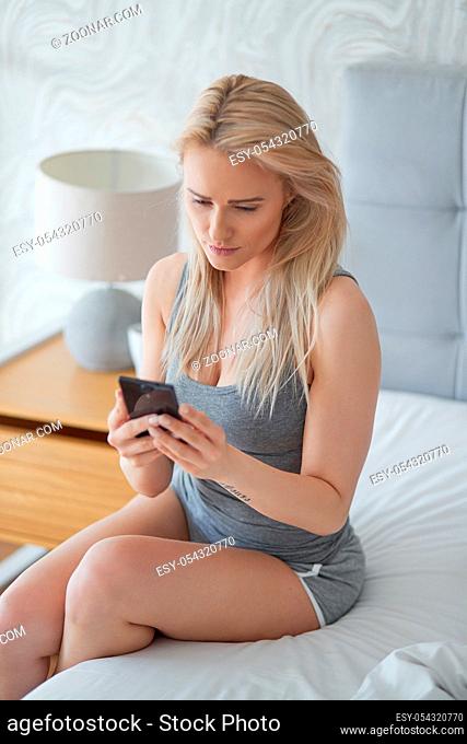 Nice middle aged woman sitting on her bed in the morning and checking smartphone for new messages. Wearing basic tank top and shorts