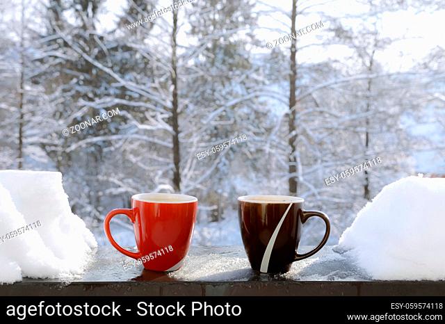 Red and brown mugs with hot drinks, on the edge of a balcony, with snow on the sides and a snowy nature in the background