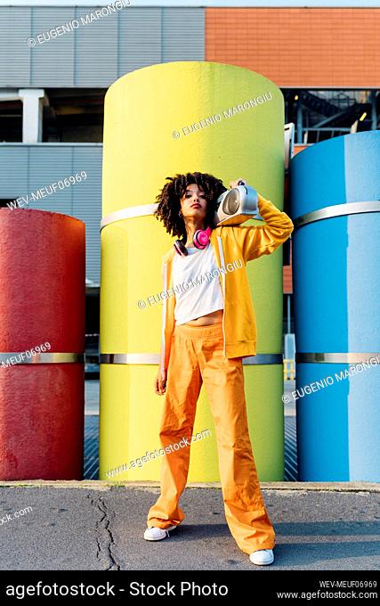 Young woman carrying boom box on shoulder in front of colorful pipes